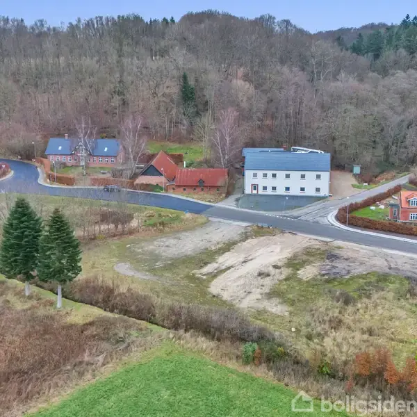 Et stort hvidt hus står ved en vej omgivet af andre huse og frodige, skovklædte bakker i baggrunden. Forgrunden viser åbne grønne marker og nogle træer.
