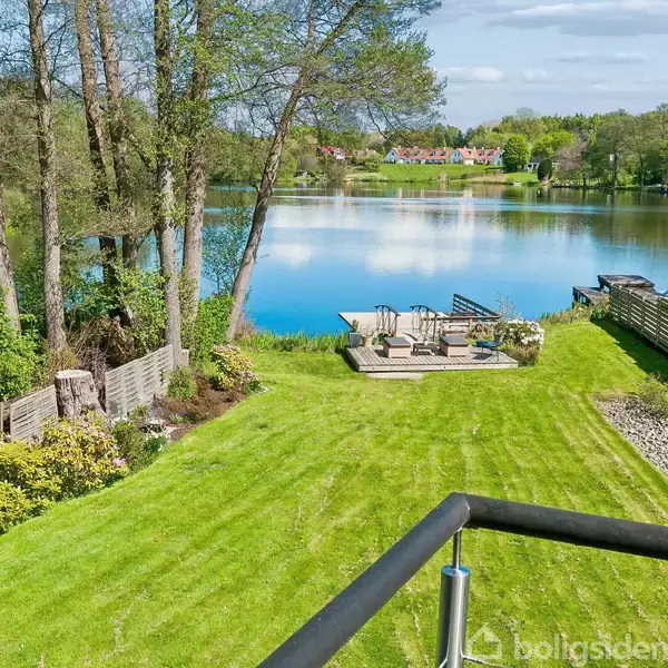 Græsplæne strækker sig ned til søbredden med terrasse og havemøbler. Træer og buske omgiver området. I baggrunden ses skov og huse på den anden side af søen.