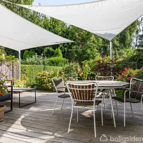 En lys stue har en beige sofa med farverige puder. Et mønstret tæppe pryder gulvet. Bag sofaen ses en spisestue med moderne stole og et stort vindue, som slipper naturen ind.