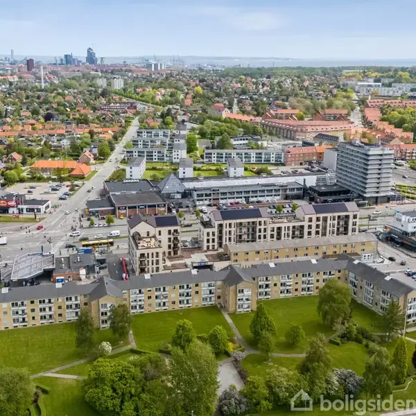 Bygninger ligger i et grønt område med træer, set fra oven. Boligblokke omkranser en parkagtig plads. Baggrunden viser en by med mange bygninger og en blå himmel.