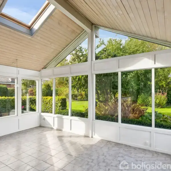 En lys havestue med flisegulv og store vinduer, der giver udsigt til en frodig have. Loftet har skrå flader med ovenlysvinduer, der slipper sollys ind.