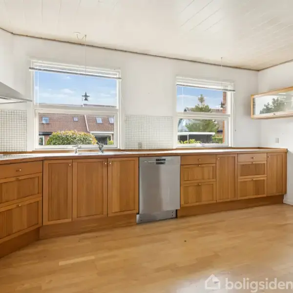 Køkken med træskabe og indbygningsovn har en opvaskemaskine i midten og et køleskab til højre. Rummet er lyst med to store vinduer og lyst trægulv.