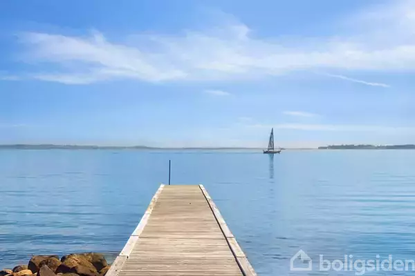 Træbro strækker sig ud over en stille sø, hvor en sejlbåd er synlig i horisonten under en klar blå himmel. Sten ligger ved bredden.