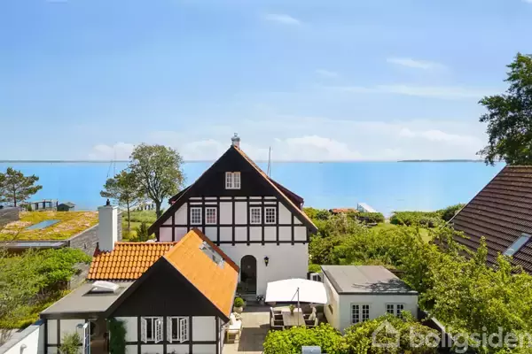 Et bindingsværkshus står stille omgivet af frodige haver og små bygninger. I baggrunden ses en rolig havudsigt under en klar blå himmel.