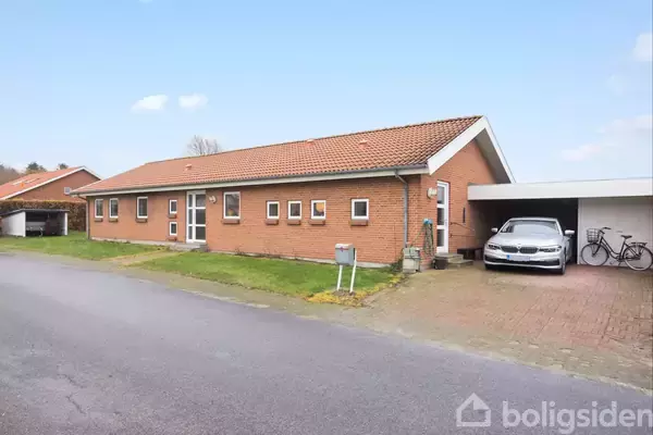 En rød murstensbungalow med carport, hvor der holder en bil og to cykler, placeret i et beboelseskvarter langs en asfalteret vej.