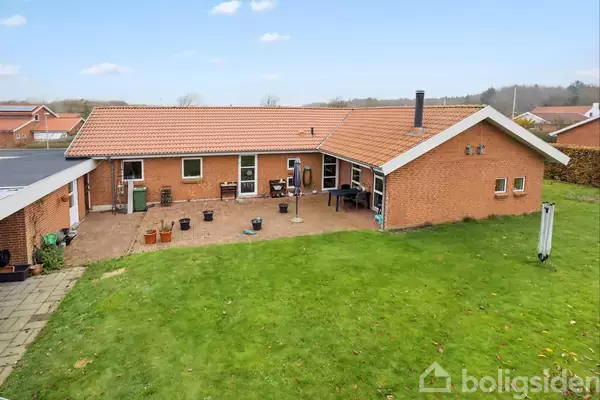 Et rødt murstenshus med terracotta-tag, står stille i en baghave omgivet af græsplæne og terrasserede områder med diverse planter.