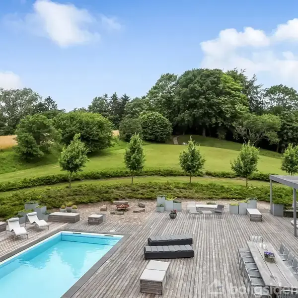 Svømmebassin står stille omgivet af en stor terrasse med loungemøbler. Grønt anlæg i baggrunden med træer og græsplæne under en blå, let skyet himmel.