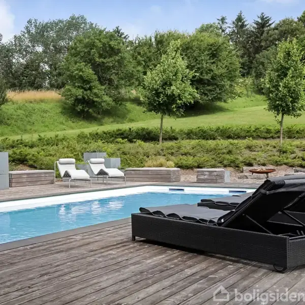 Sort lounge møbler står ved en rektangulær swimmingpool på en træterrasse. I baggrunden ses en græsplæne med buske og træer, hvilket skaber en idyllisk udendørs atmosfære.