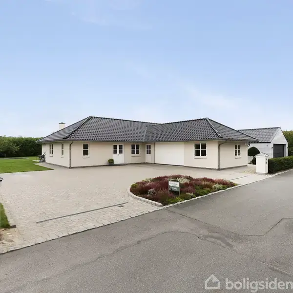 En hvid bungalow med mørkt tag står ved en bred indkørsel. Den er omkranset af en velplejet have. Husnummeret "10" ses på en postkasse ved indkørslen.