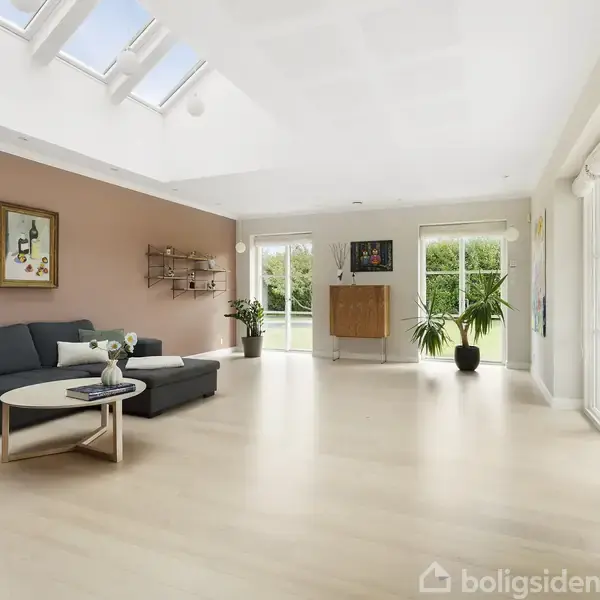 Stuen har en sort sofa med et rundt bord, belyst af ovenlys. Væggen er lys rosa med et maleri. Store vinduer åbner op til en grøn have.