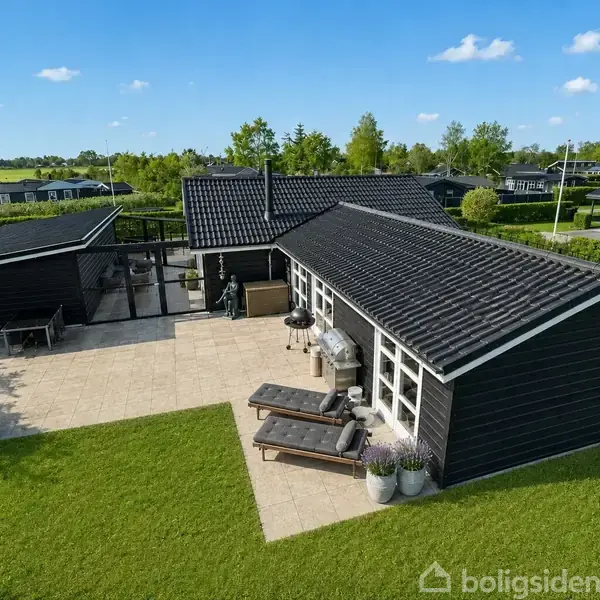 Sort træhus med terrasse i solen, omgivet af grøn plæne. Terrassen har liggestole og planter. Flere huse ses i baggrunden under en blå himmel med spredte skyer.