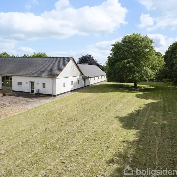 Murstenshus med sort tag står stille i en grøn have med træer og buske. Græsplænen er velholdt, og der er en terrasse med havemøbler foran huset.