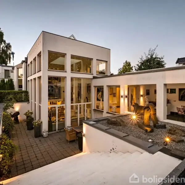 Moderne hvid villa med store vinduer, omgivet af en flisebelagt gårdhave. Haven har skulpturer og dekorative planter, mens det indre er oplyst og synligt gennem de glasfacader.