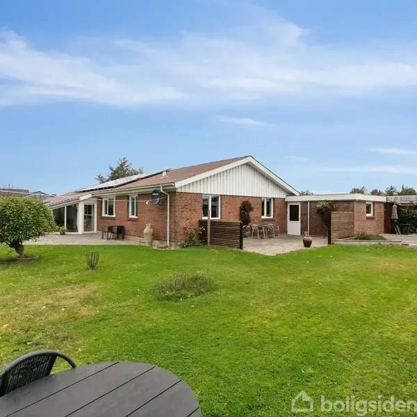 En murstensbygning med saddeltag står i en grøn have med klippet græsplæne. Der er et rundt bord og stole i forgrunden, omgivet af en åben, klar himmel.