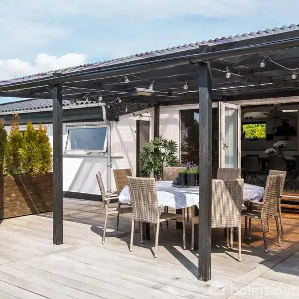 Udendørs terrasse med kurvemøbler, spisearrangement under tag, omgivet af grønne planter. Store vinduer åbner ind til en moderne stue. Himlen er klar og solrig.