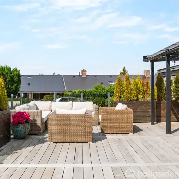 Terrasse med flettede møbler og borde under en halvtag. På trædækket er der blomsterkrukker og planter. Baggrunden viser nabohuse og en blå himmel.