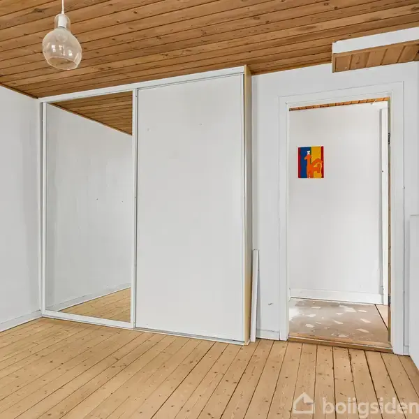 Et tomt værelse med trægulv og træloft. Skydedørsskabet er lukket. En enkel lampe hænger fra loftet. Døren fører til en gang, hvor et lille farverigt maleri ses på væggen.