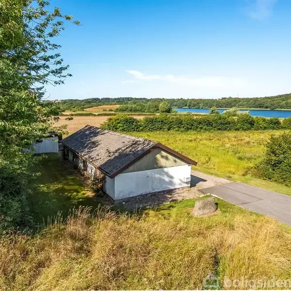 Et hus står stille omgivet af frodige marker og en sø i baggrunden under en klar blå himmel. En vej løber langs husets ene side.