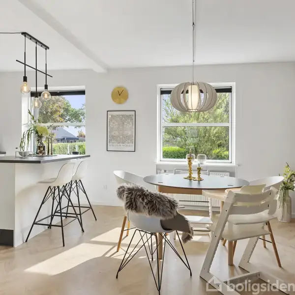 Et moderne køkkenalrum med en hvid køkkenø og sorte stole, ved siden af et spisebord med hvide stole. Store vinduer giver udsigt til grøn natur udenfor. En gul vægur og tre vægkunstværker pryde væggen.