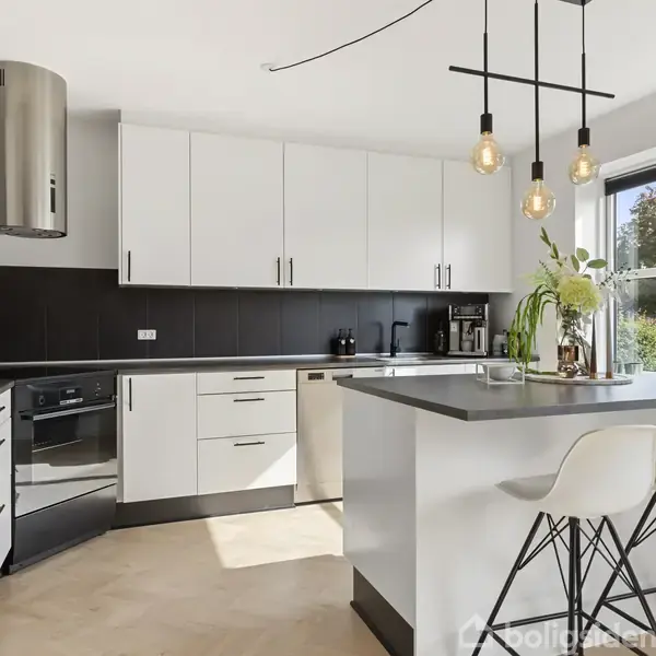 Køkken med hvide skabe og sort bordplade, indeholdende en ovn og opvaskemaskine. To hvide stole står ved en køkkenø, og store vinduer giver udsigt til grønne træer udenfor.