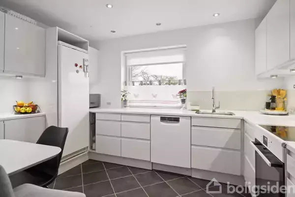 Et moderne køkken med hvide skabe og stålgreb, integreret opvaskemaskine og komfur. Et stort vindue over køkkenbordet, der giver lys. Frugtskål og kaffemaskine på bordet.