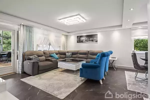 En stor hjørnesofa står i en lys stue med et moderne design. Et sofabord er centralt placeret, omgivet af blå stole. Store vinduer giver udsigt til en terrasse.