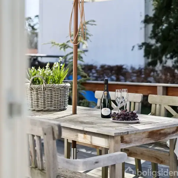 Træbord med vinflaske, to glas, en kurv med blomster og en klase druer. Bordet står på en terrasse med grønne omgivelser og en hvid bygning i baggrunden.