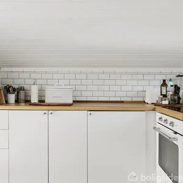 Et køkken med hvide skabe og en brødrister på bordpladen. Der er flisevægge, skærebrætter, glasbeholdere, køkkenredskaber og olier. En ovn er indbygget i skabet.
