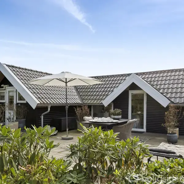 Et sortmalet hus med skråtag står i haven. Foran huset er en terrasse med spisebord, stole og en parasol. Omgivet af grønne buske under en klar blå himmel.