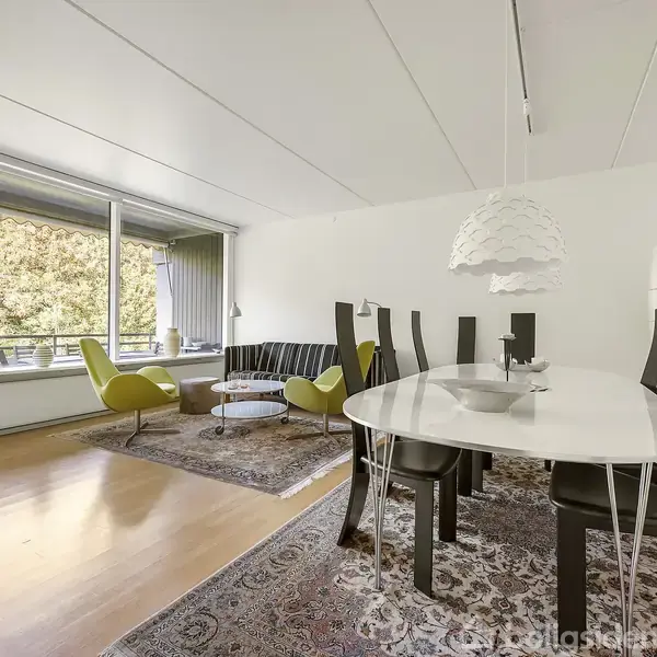 Et moderne, stilfuldt spisebord med sorte stole står på et tæppe. I baggrunden er en stue med en stor sofa, to grønne stole og store vinduer, der giver udsigt til træer.