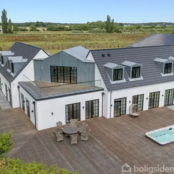 Et moderne, hvidt hus med mange vinduer står på en stor træterrasse. Terrassen har møbler og et spabad. Omgivet af grønt område og marker i baggrunden.