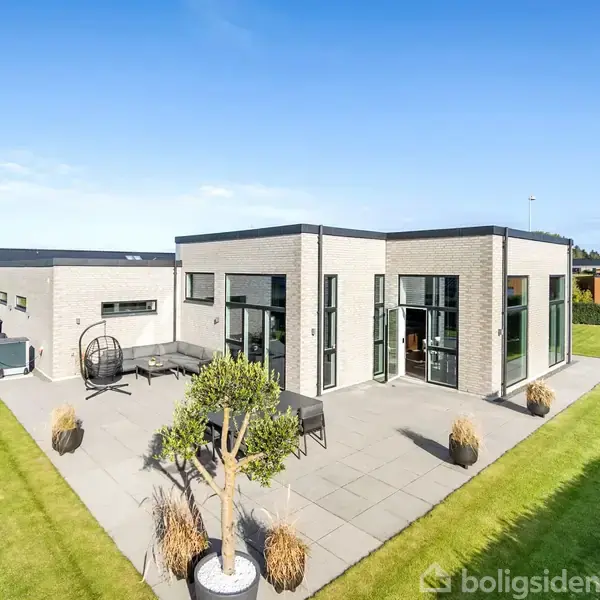 Modern villa med fladt tag har store vinduer og er omgivet af en velplejet have og stenlagt terrasse med havemøbler og planter. Blå himmel i baggrunden.
