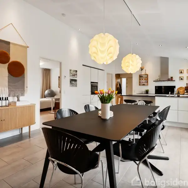 Et moderne spisekøkken med sort spisebord og sorte stole i midten. Lyse lamper hænger over bordet. Rummets vægge er hvide, med et skab fyldt med dekorationer til venstre.