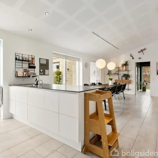 Et moderne køkken-alrum har et hvidt køkkenbord i forgrunden, med barstole ved siden. Til venstre ses en terrasse med parasol gennem en skydedør. Loftslygter lyser rummet.