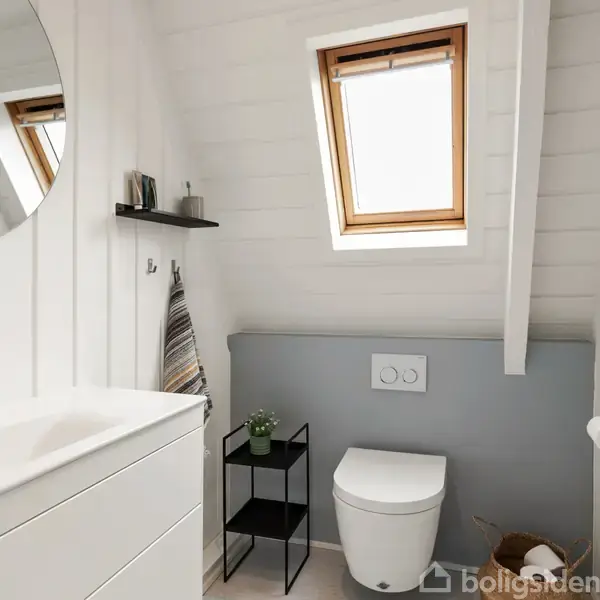 Rundt spejl hænger over en hvid vask på et badeværelse. Toilet placeret ved skråvæg med ovenlysvindue. Sort hylde og plante i en minimalistisk indretning. Rolig atmosfære.