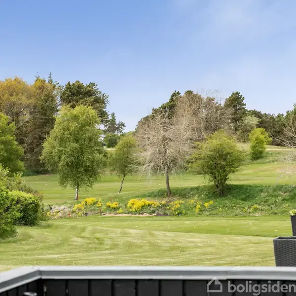 En grøn plæne omkranset af spredte træer strækker sig ud fra en terrasse med sorte møbler. Solrigt vejr og en klar, blå himmel danner baggrund.