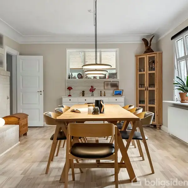 Et spisebord med otte stole står i et lyst rum. Der er en pejs tændt, en grøn skænk ved et vindue, og en reol er fyldt med genstande.