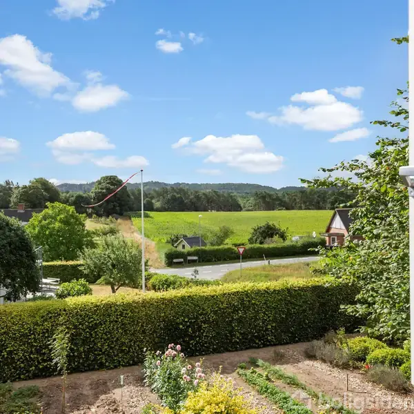 Åben vindue med udsigt over en velholdt have med blomsterbede i forgrunden. En lav hæk skiller haven fra grønne marker og skov i baggrunden under en klar blå himmel.