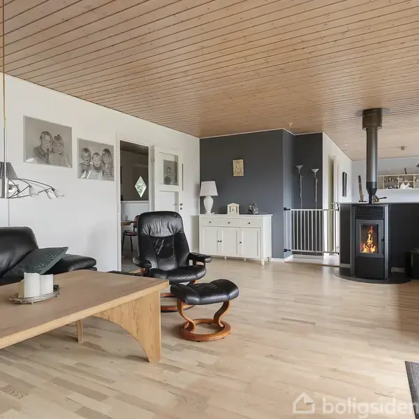 Sort sofa og lænestol står ved et træbord i en stue med mørkegrå vægge og brændeovn, omgivet af trægulv, hvide paneler og store vinduer.