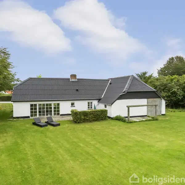 Hvidt hus med sort tag står på en grøn græsplæne. To liggestole er placeret foran huset. Omkring er der tæt bevoksning af træer og buske.