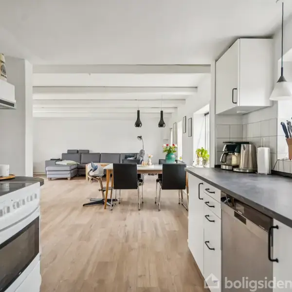 Køkken med hvid bordplade, kaffemaskine og komfur. Køkkenet hænger sammen med en åbent spisestue med spisebord og stole. Lys strømmer ind gennem store vinduer.