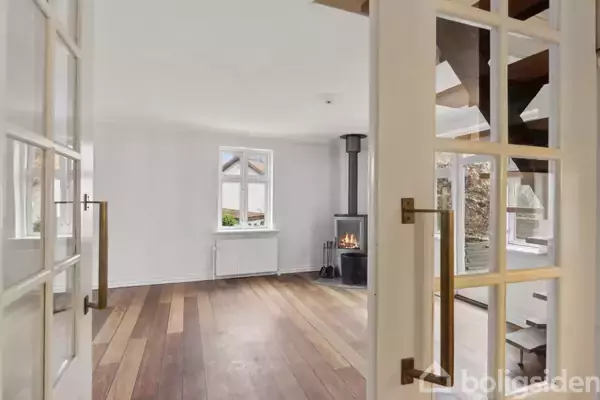 Franske døre åbner op til en stue med trægulv. Stuen har en brændeovn i hjørnet og hvidmalede vægge. Vinduer slipper naturligt lys ind.