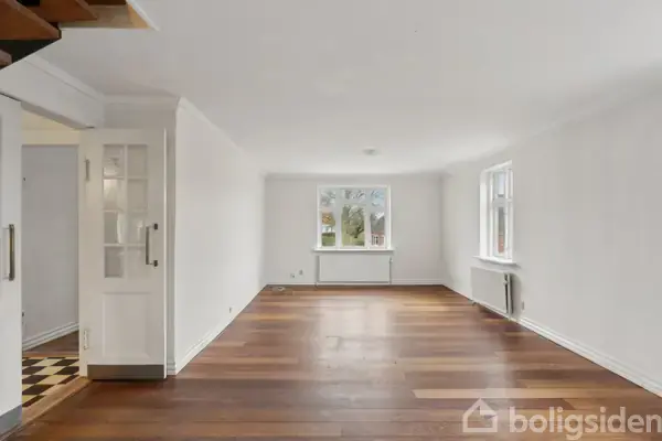 En rummelig stue med trægulv og hvide vægge, indeholder en åben dør til venstre. To vinduer giver udsigt udadtil, og rummet er lyst og tomt.