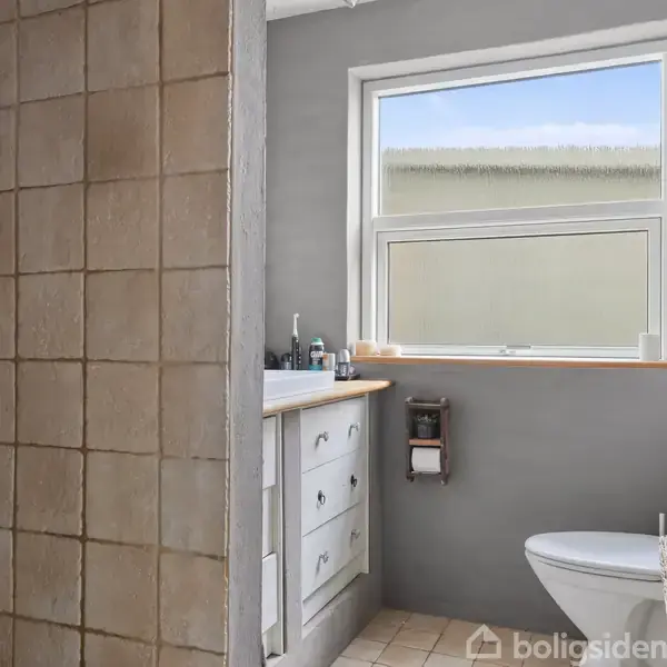 Badeværelse med flisebelagt brusekabine, vask med skuffer, spejl, og vindue. Toilet ved siden af vasken. Hylde med toiletartikler hænger på brusevæggen.