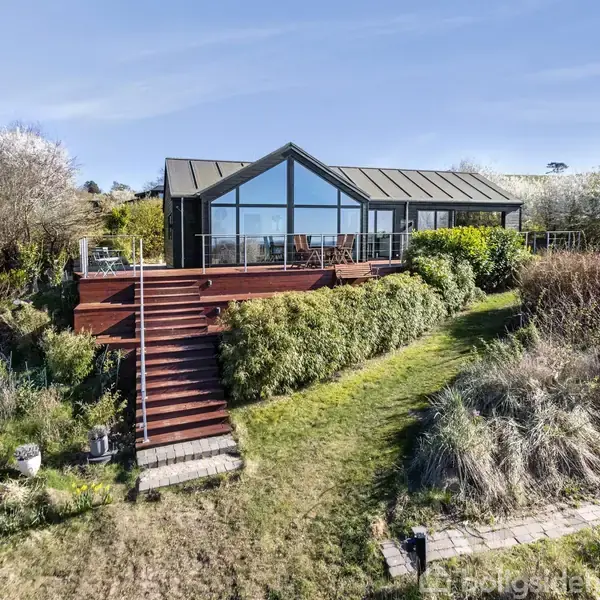 Et moderne hus med store glaspartier står på en hævet træterrasse. Terrassen er forbundet til en trappe ned til en frodig have med tæt beplantning og græsplæne.
