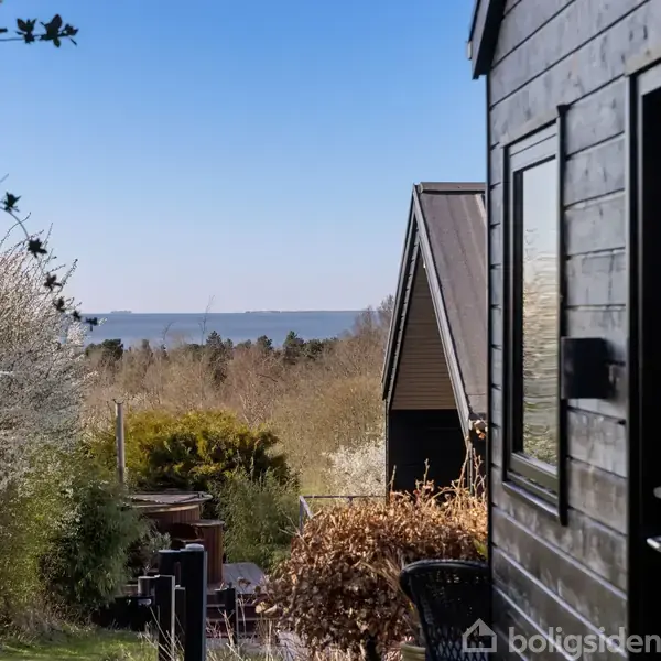 Sort træhus med vinduer står ved siden af blomstrende buske. I baggrunden ses havet under en klar blå himmel, omgivet af træer og grønne områder.