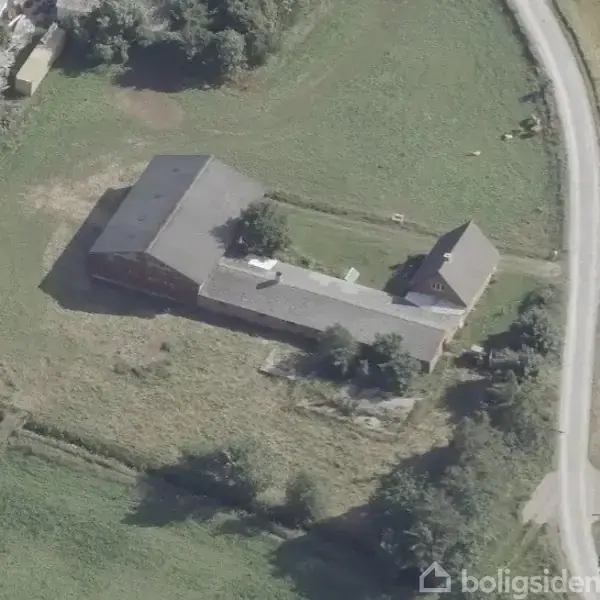 Bygning ligger i en landlig omgivelser, flankeret af grusveje og grønne marker. Tre bygninger danner en vinkel, og spredte træer ses omkring strukturen.