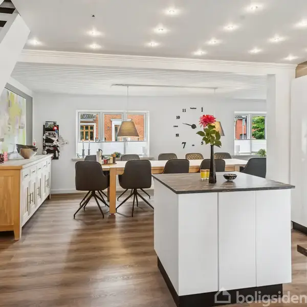 Køkken-alrum med hvidt interiør og trægulv rummer et spisebord til seks personer med udsigt til vægdekoration med ur. En trappe leder til en øvre etage.