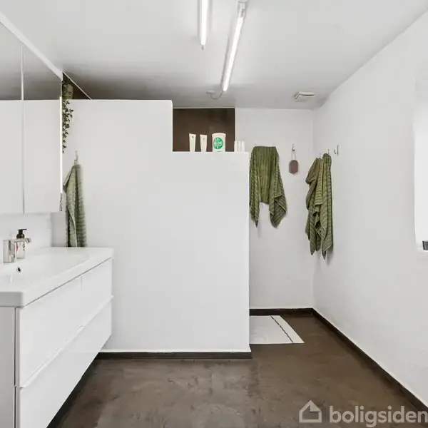 Håndvask med to skuffer står ved væggen, omgivet af hvide vægge og grønne håndklæder hængende ved siden af. Tredelt spejl ovenover, med fliser på gulvet.