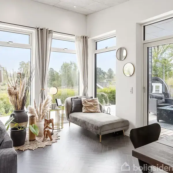 Stue med grå sofa og liggestol, udsigt gennem store vinduer til have. Dekoreret med planter, lamper og puder. Bord til højre, spejle på væggen, terrassedør åben til udendørsområde.